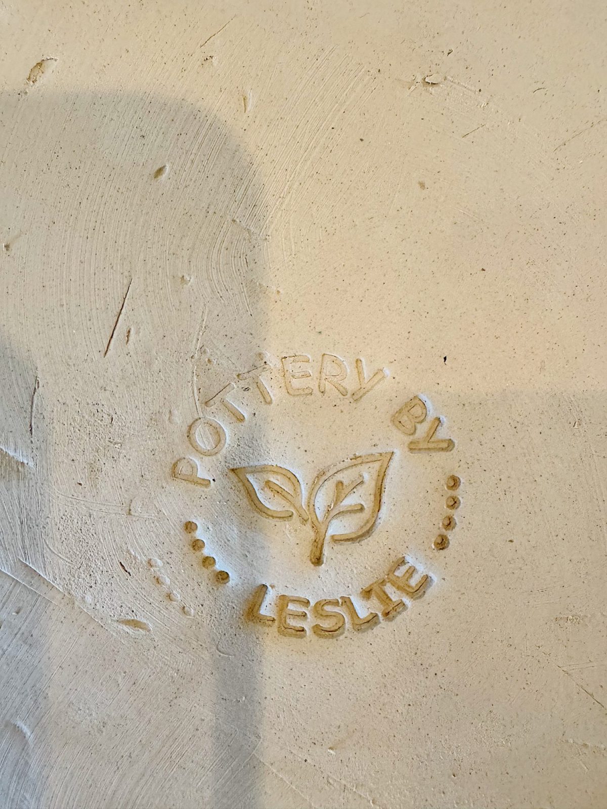 A clay surface stamped with the words &ldquo;Pottery by Leslie&rdquo; around a simple line drawing of two leaves.