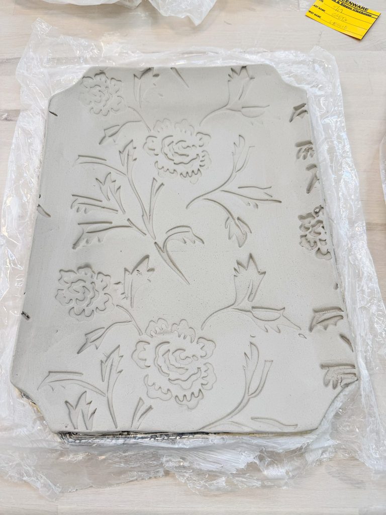 A rectangular clay tray with embossed floral patterns rests on a sheet of plastic wrap on a light-colored surface.