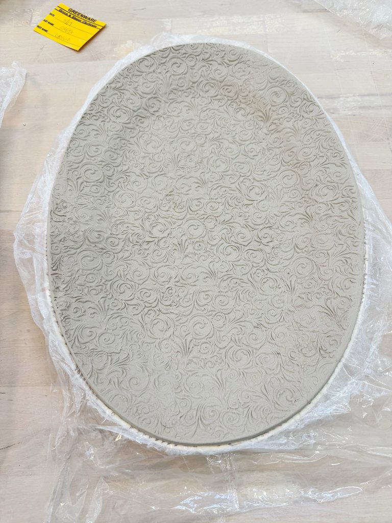 An oval ceramic plate with an intricate, raised swirl pattern rests on clear plastic wrap atop a light wooden surface.