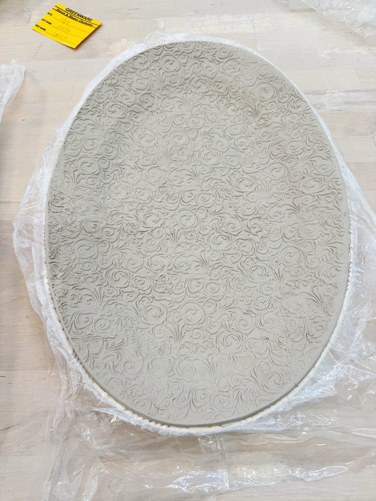An oval ceramic plate with an intricate, raised swirl pattern rests on clear plastic wrap atop a light wooden surface.