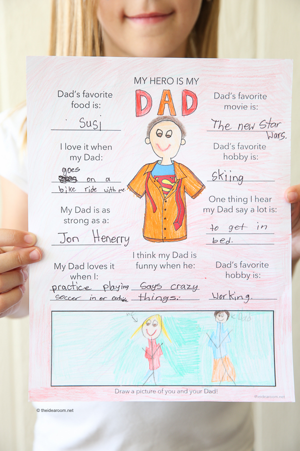 A child holds a hand-drawn and colored worksheet titled "My Hero Is My Dad," featuring answers about their dad's favorites, hobbies, and a drawing of them together at the bottom.