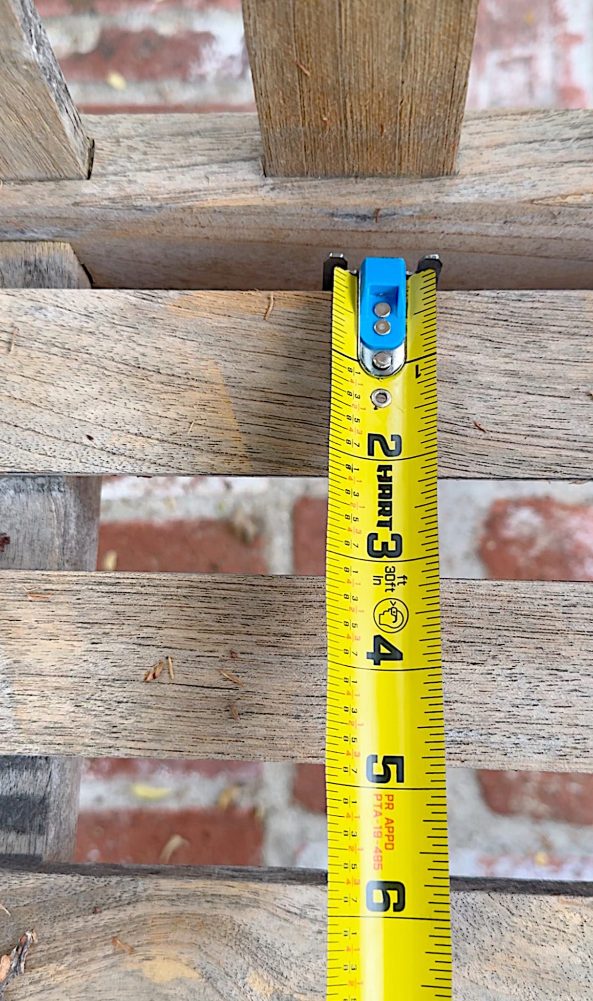 A yellow tape measure extended to about 4.5 inches, placed across wooden slats with brickwork visible in the background.