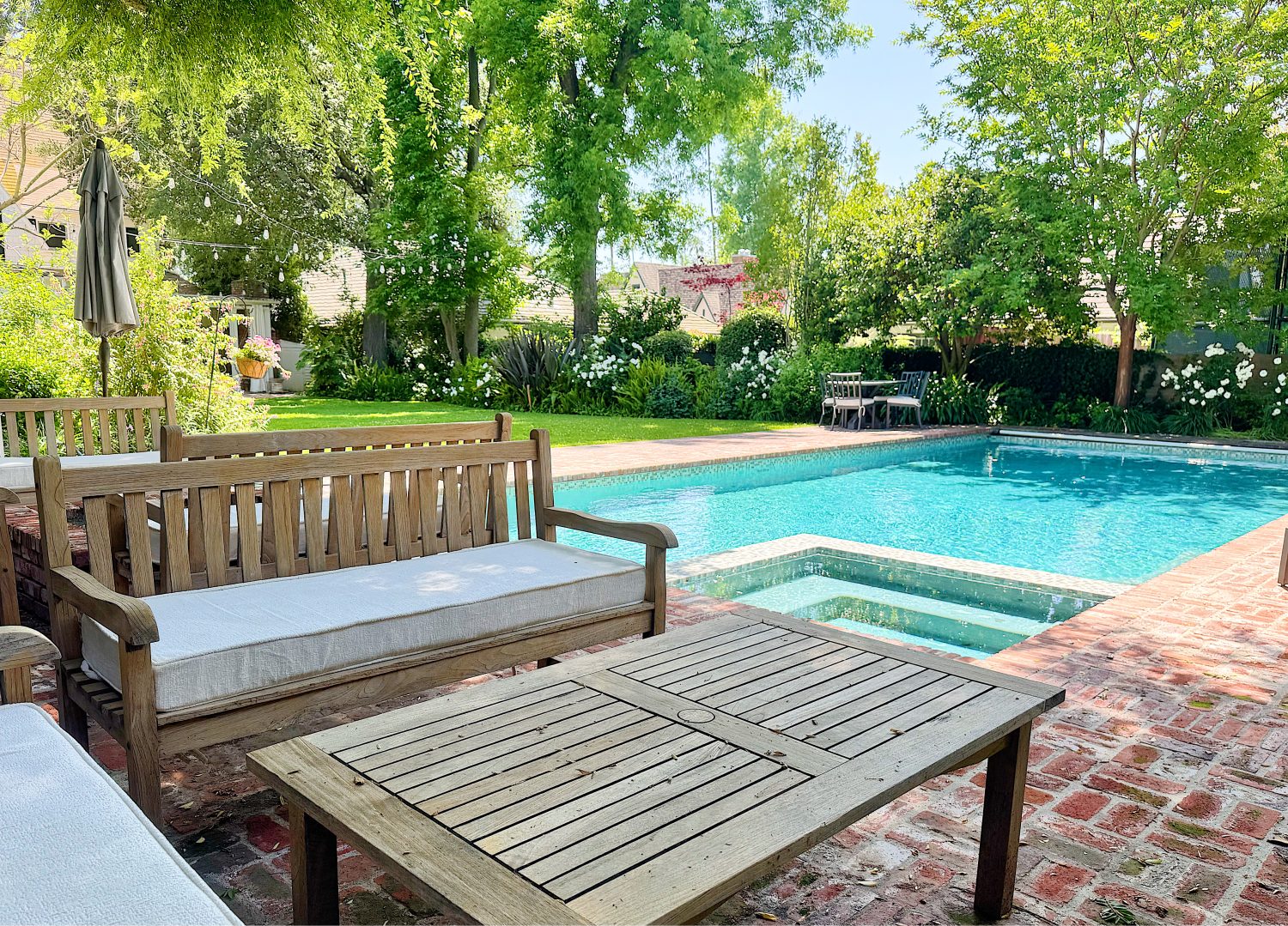 Wooden patio furniture sits on a brick patio beside a rectangular swimming pool surrounded by green trees and a landscaped garden.