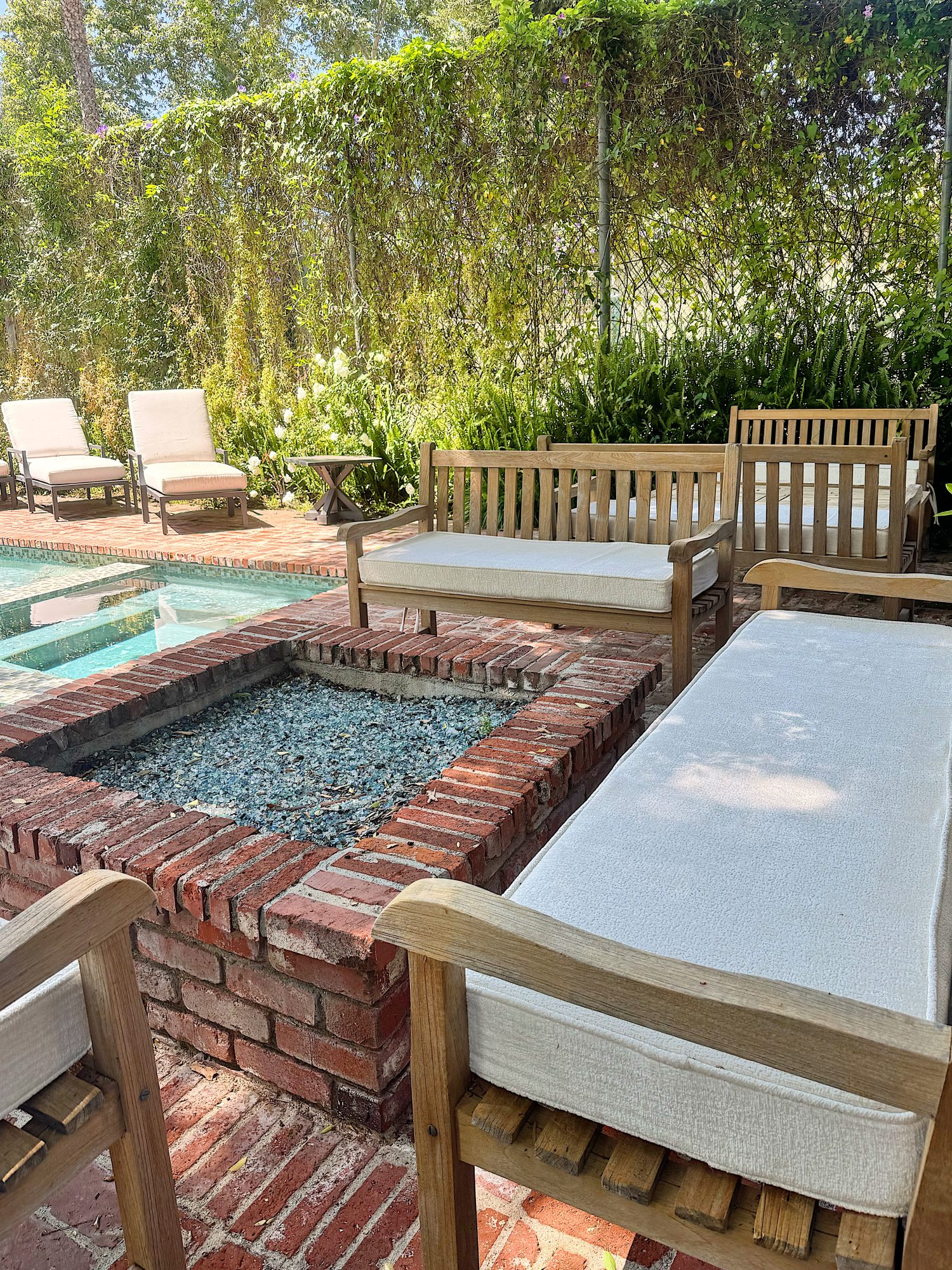 Outdoor patio with wooden furniture and white cushions arranged around a brick fire pit near a swimming pool, surrounded by greenery and plants.