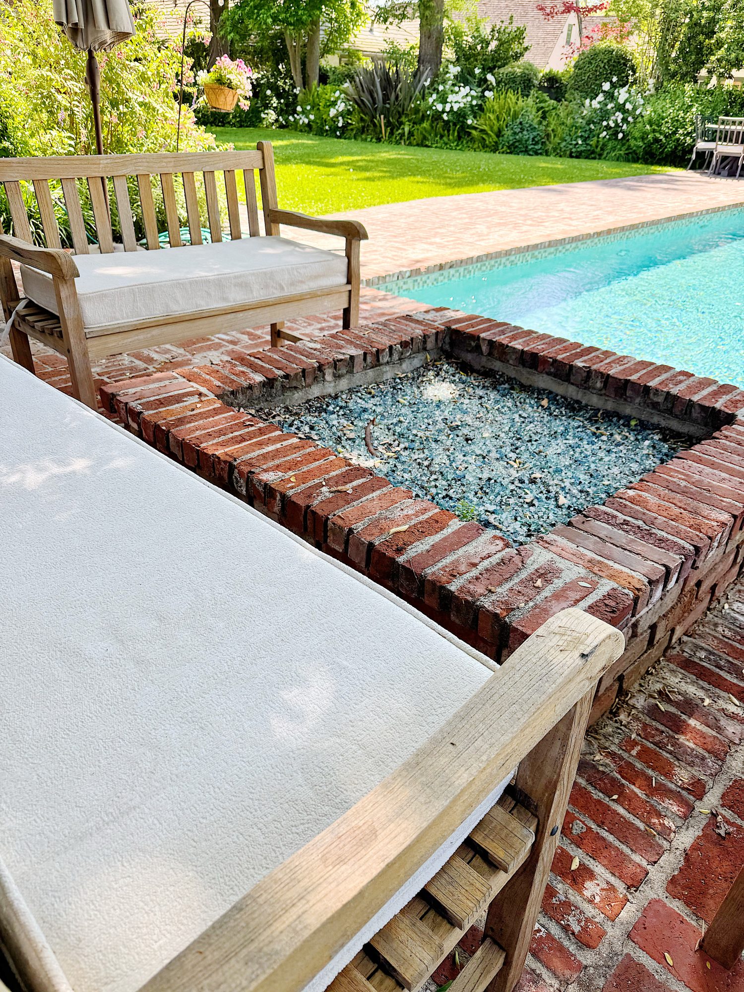 Wooden benches with white cushions surround a brick fire pit filled with glass stones, next to a swimming pool in a landscaped backyard.