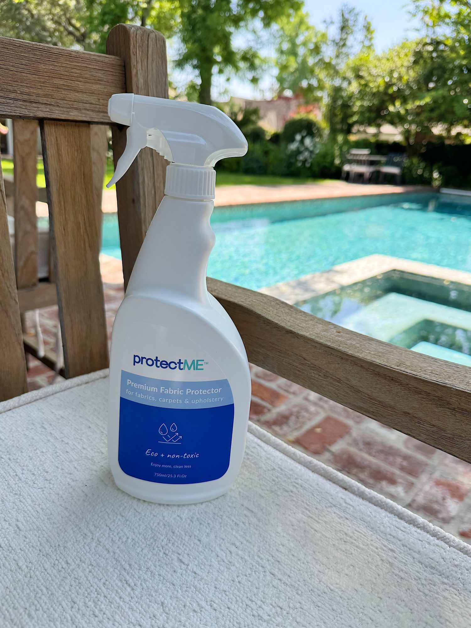 A bottle of protectME Premium Fabric Protector sits on a white chair by a pool, with trees and a brick patio in the background.