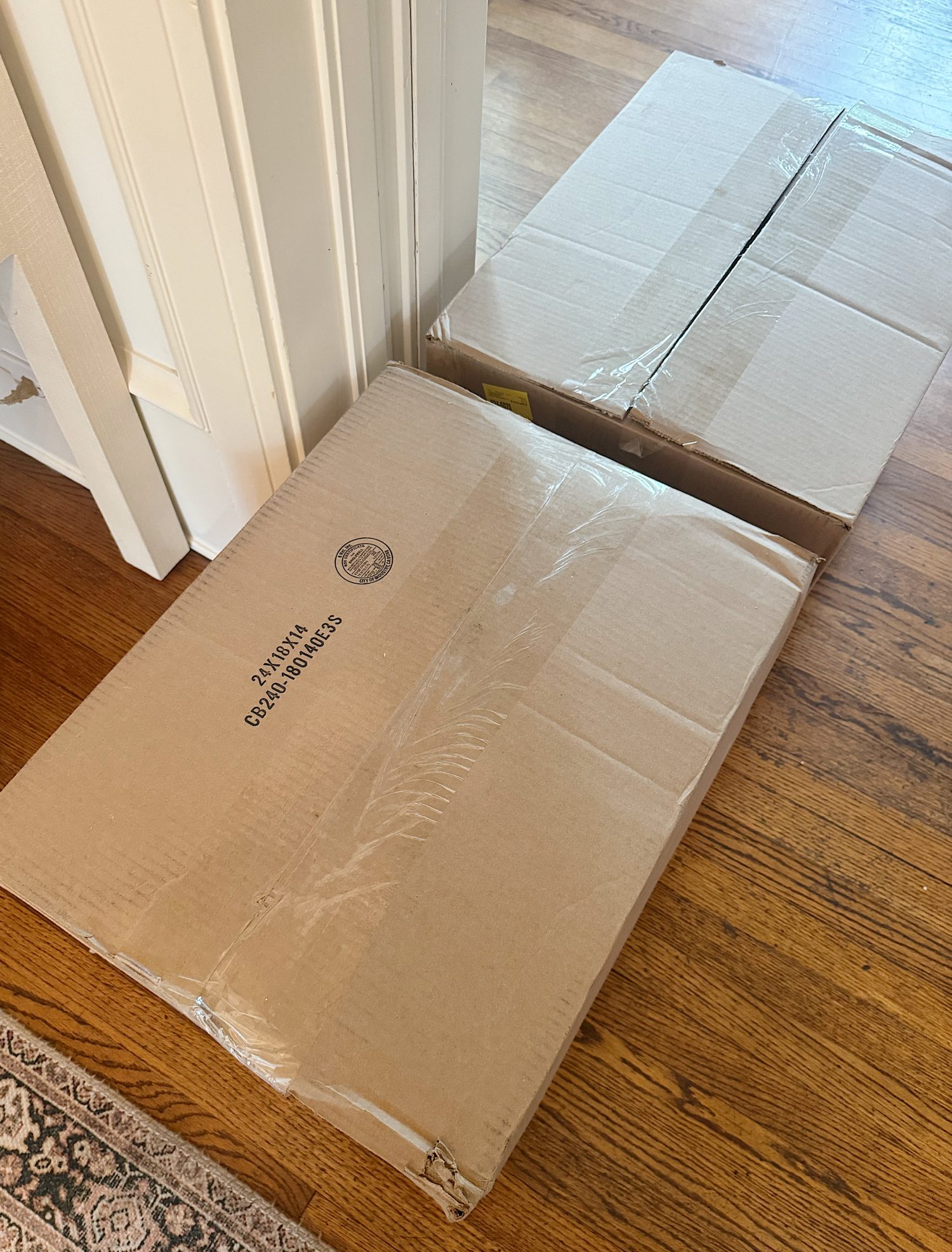 Two large closed cardboard boxes sit on a hardwood floor near a white door and a patterned rug.