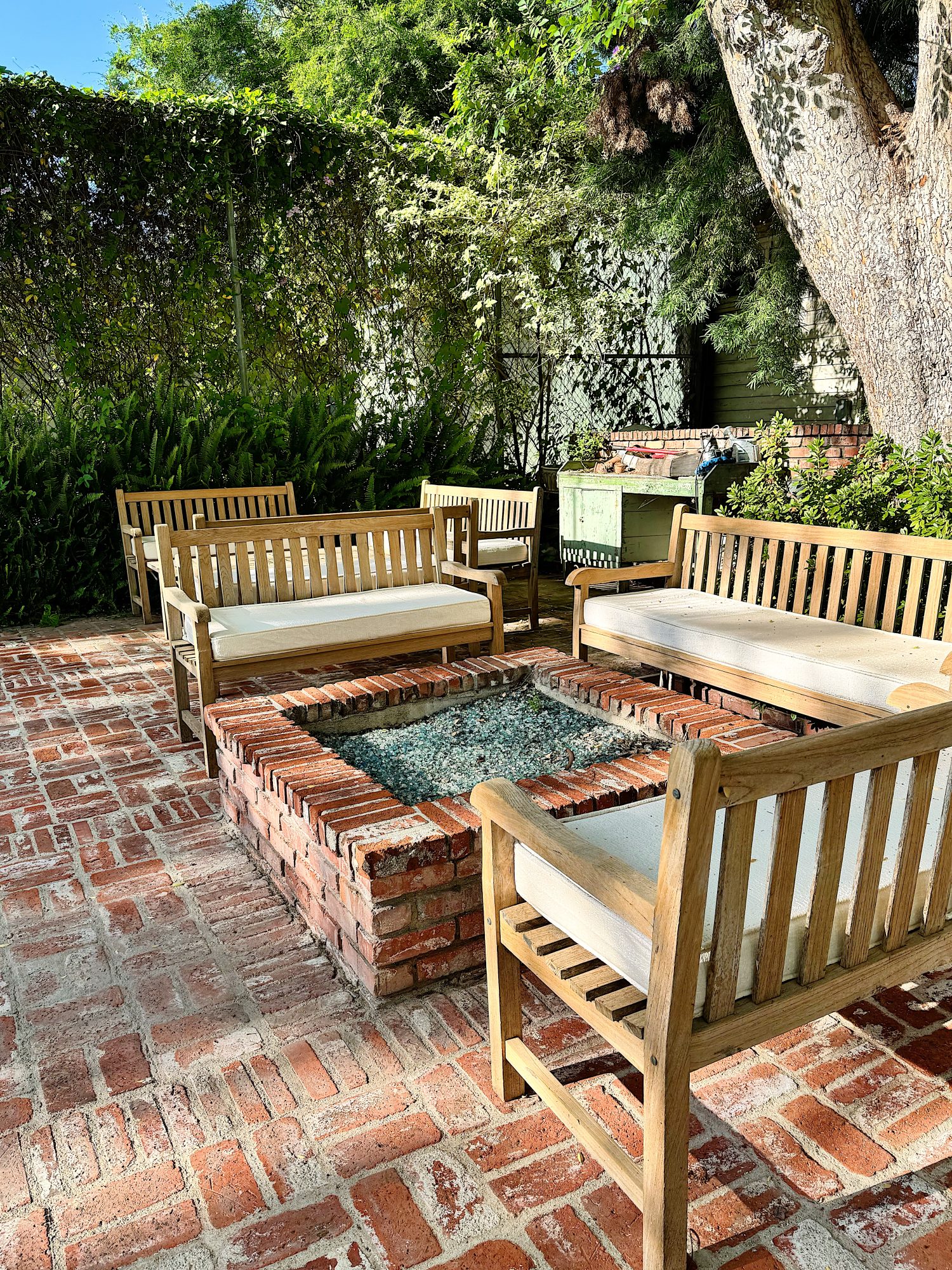 Four wooden benches with white cushions surround a rectangular brick fire pit on a red brick patio, shaded by trees and greenery.