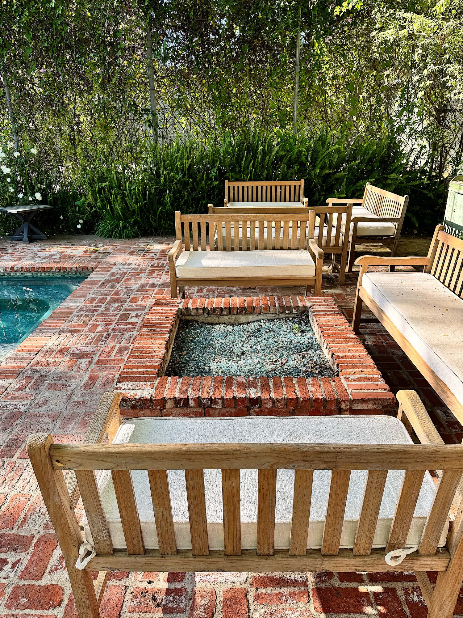 Wooden patio furniture surrounds a brick fire pit on a red brick patio next to a swimming pool, with greenery in the background.