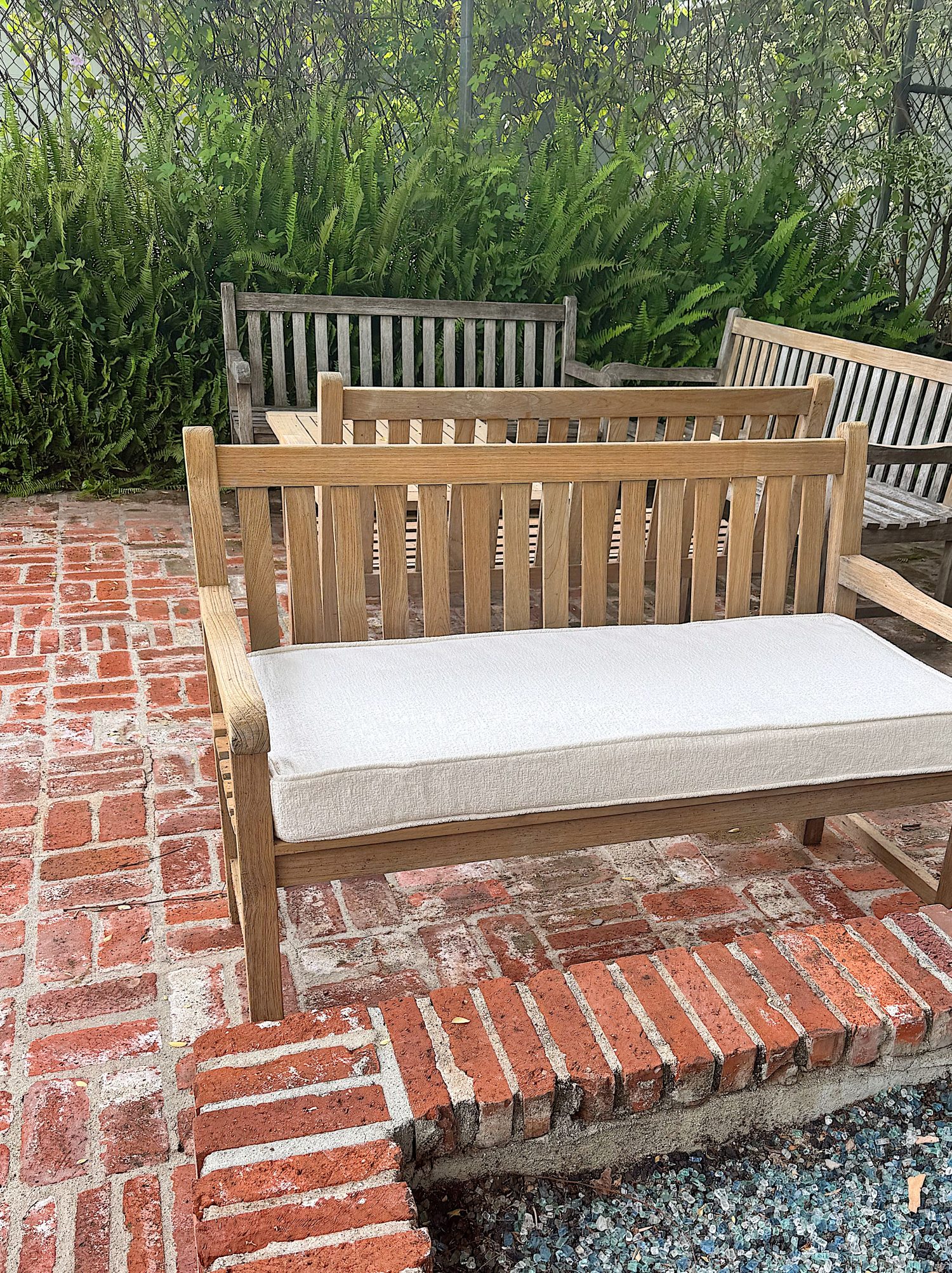 Wooden benches with slatted backs are arranged on a brick patio, with ferns and greenery in the background. The front bench has a white seat cushion.