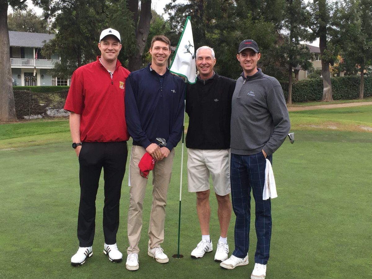 Four men stand on a golf course green near a flag, smiling at the camera. Trees and buildings are visible in the background.