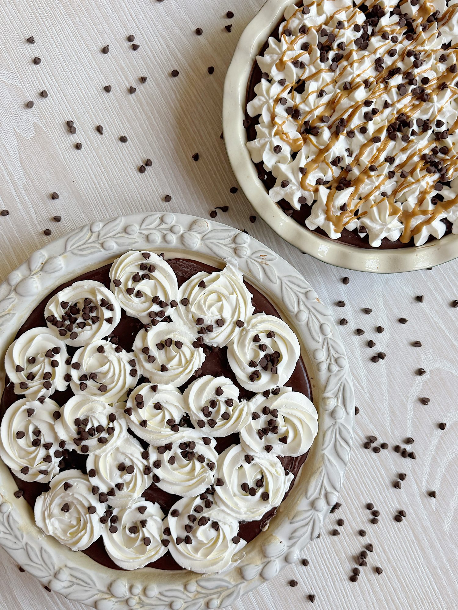 Two chocolate cream pies with whipped cream and chocolate chips on top, one with caramel drizzle, on a light surface sprinkled with chocolate chips.