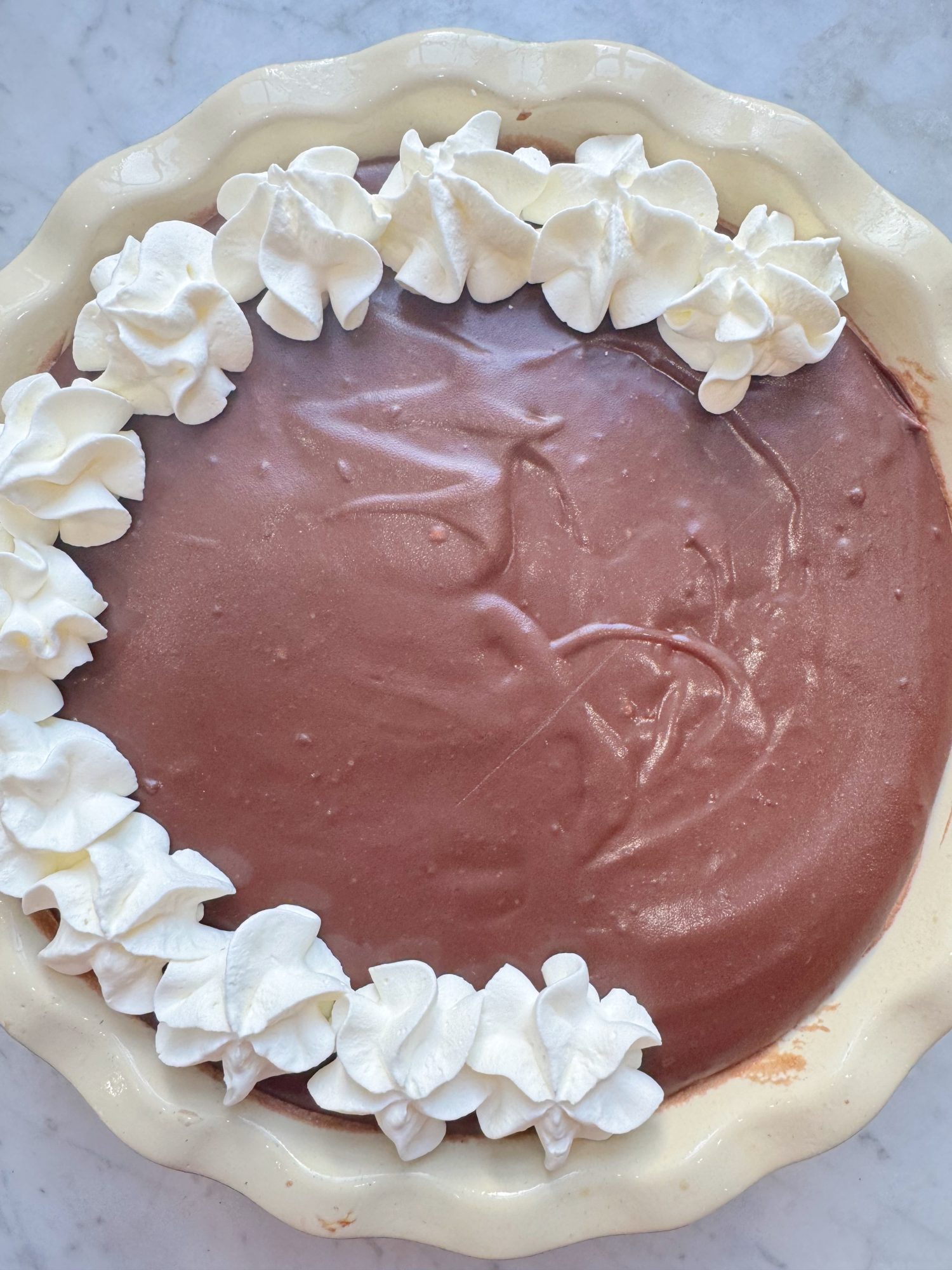 Chocolate cream pie in a pie dish topped with a border of whipped cream on one side, placed on a marble surface.