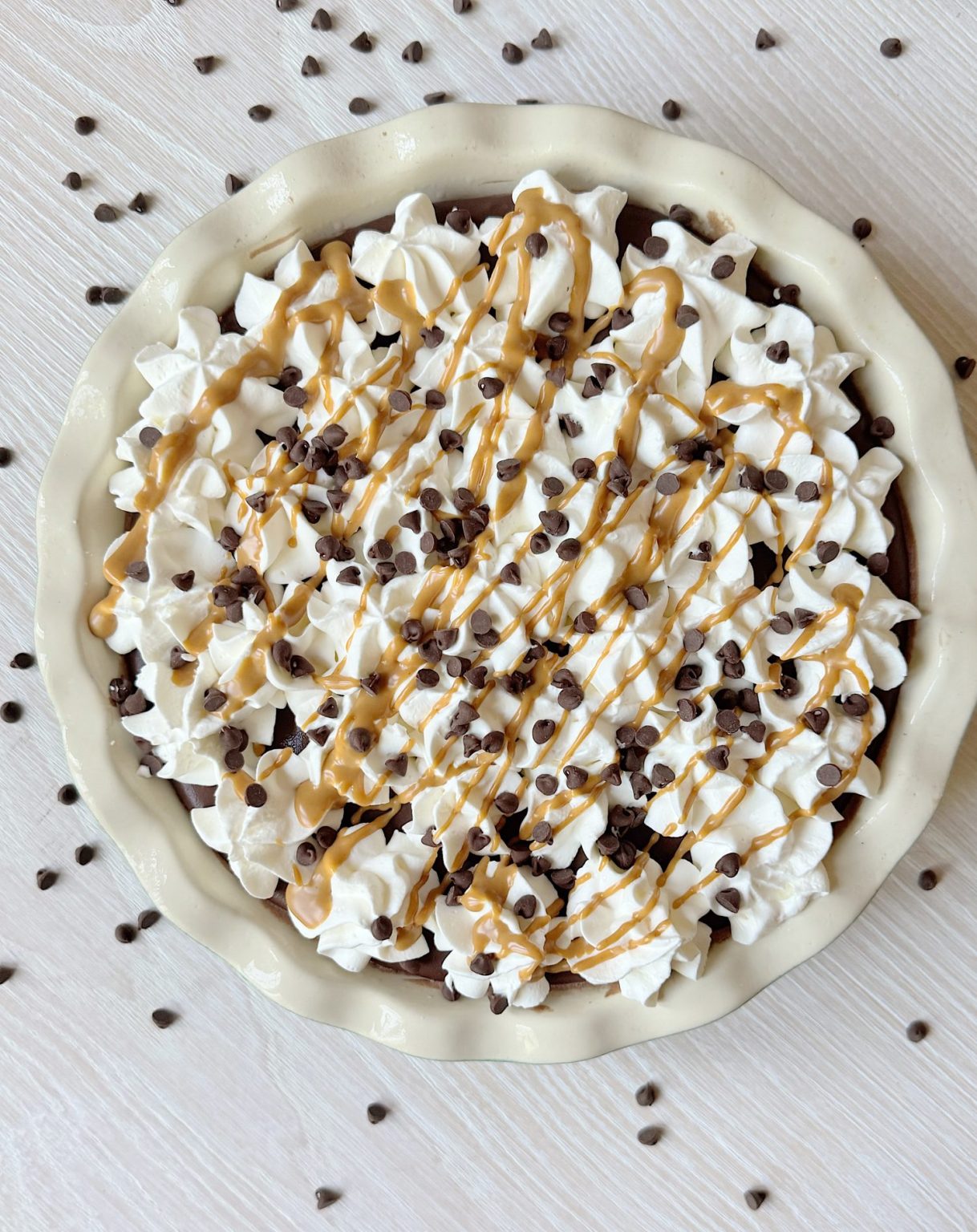 A pie topped with whipped cream, mini chocolate chips, and a drizzle of caramel, shown in a white dish on a light-colored surface with scattered chocolate chips.