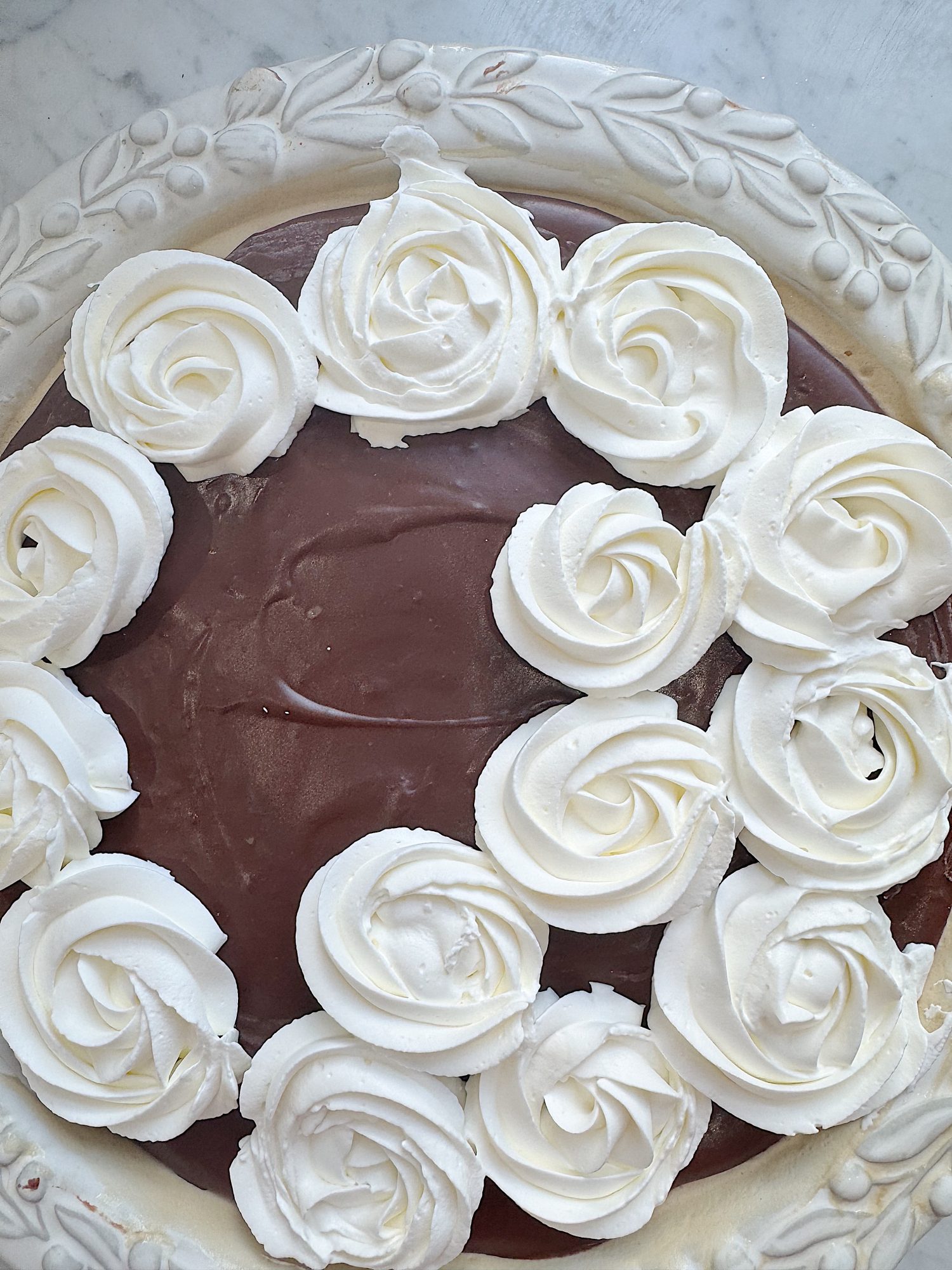 A chocolate cake topped with white whipped cream rosettes, arranged around the edge, on a decorative white plate.