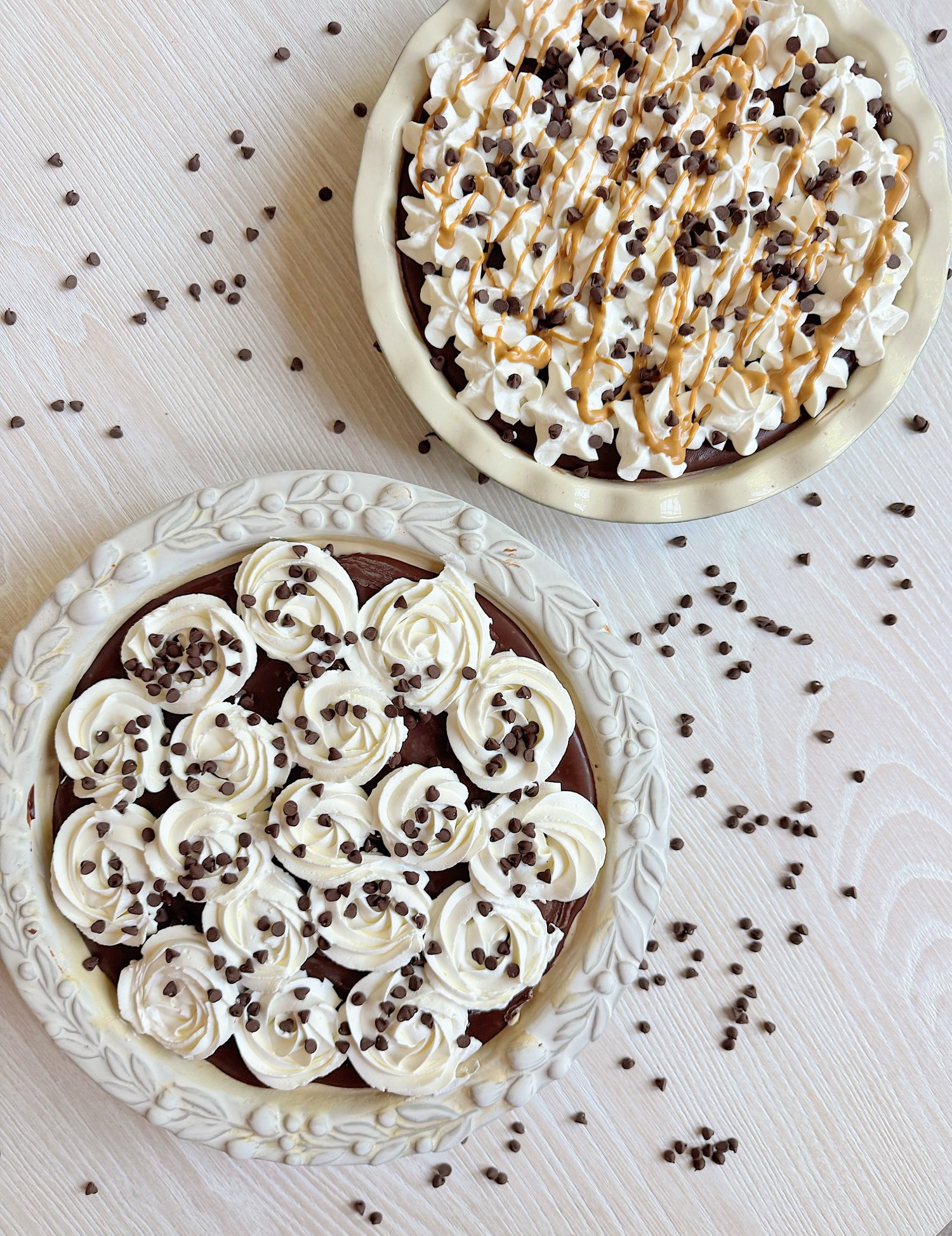 Two chocolate cream pies topped with whipped cream and chocolate chips on a light wooden surface, with chocolate chips scattered around.