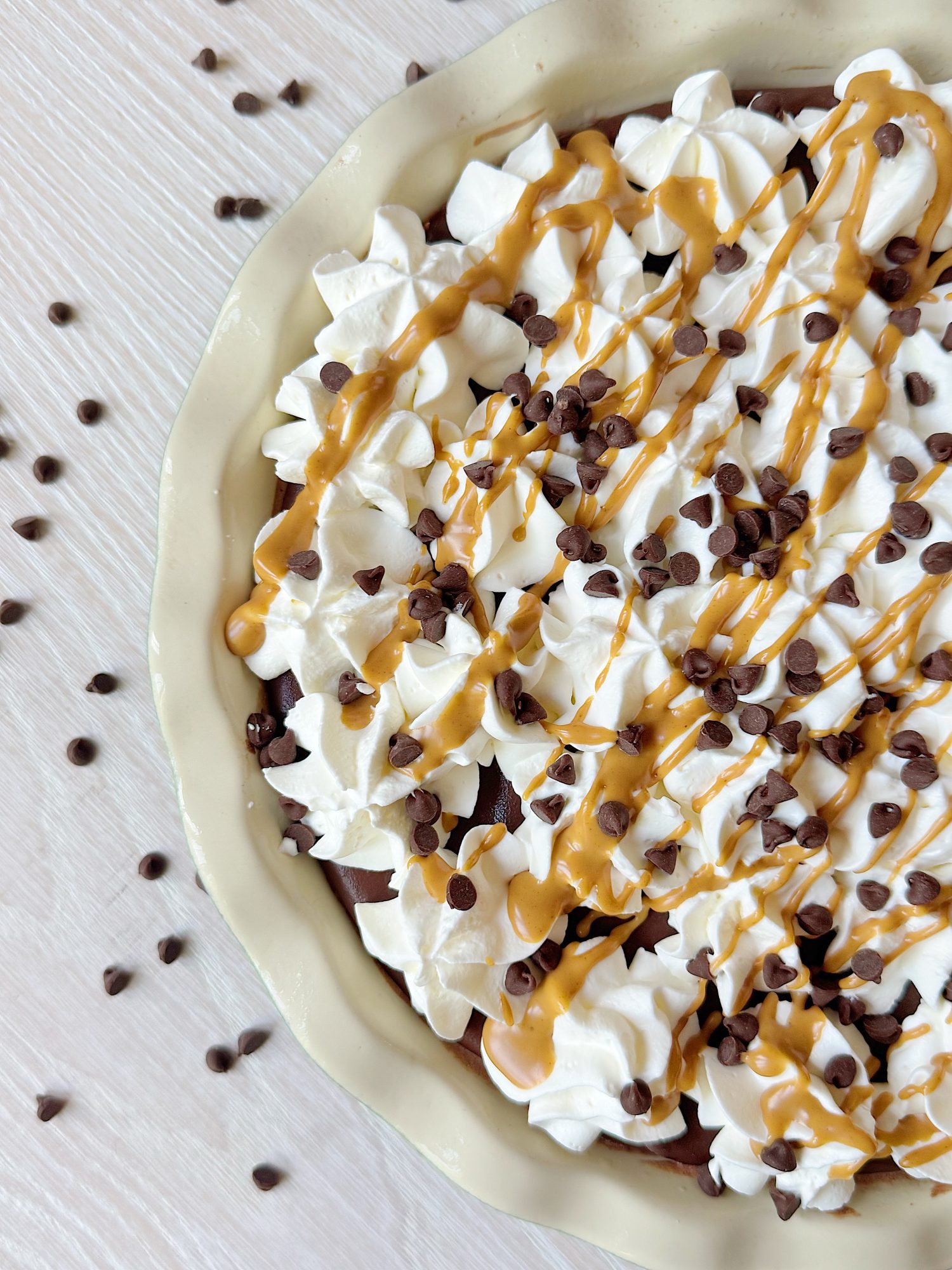 A pie topped with whipped cream, chocolate chips, and drizzled peanut butter, in a ceramic dish, with chocolate chips scattered on the light-colored surface.