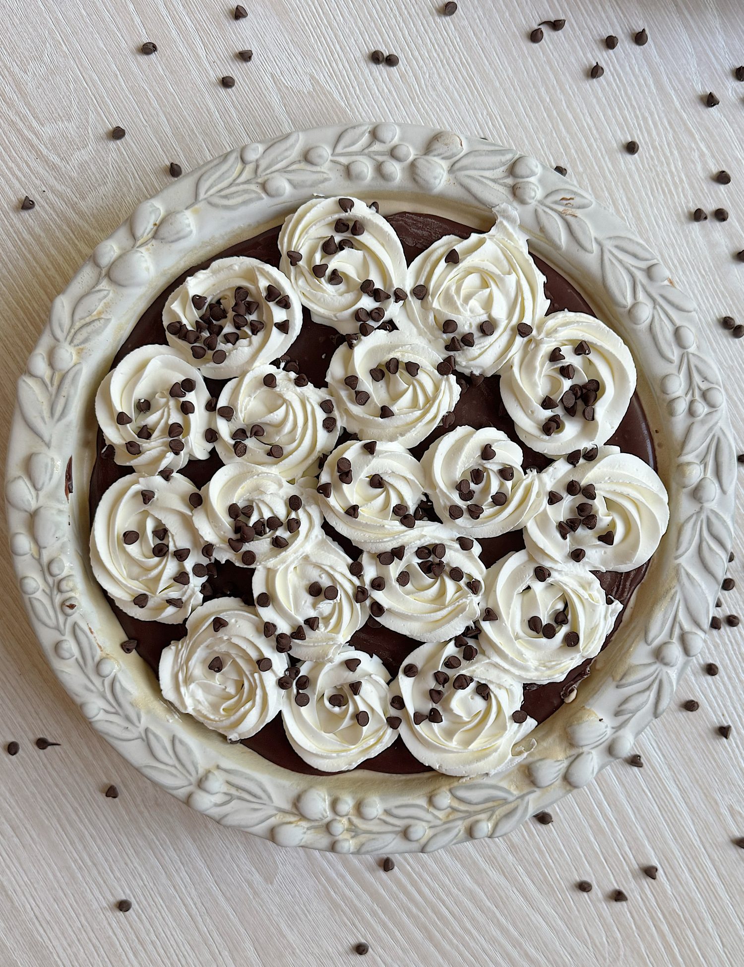 A chocolate pie topped with swirls of whipped cream and sprinkled with chocolate chips, presented in a decorative pie dish on a light wooden surface.