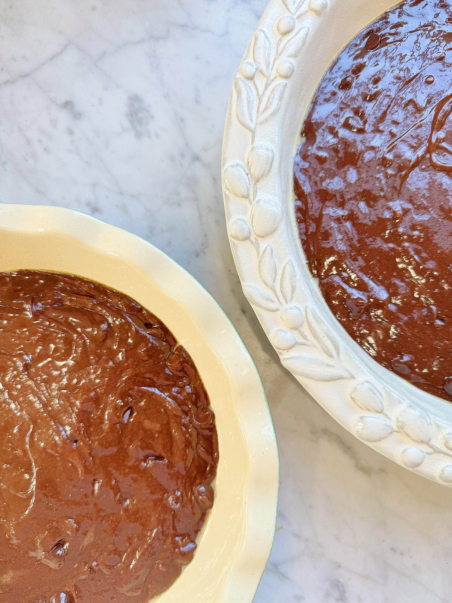 Two unbaked chocolate pies in ceramic pie dishes on a marble surface, viewed from above.