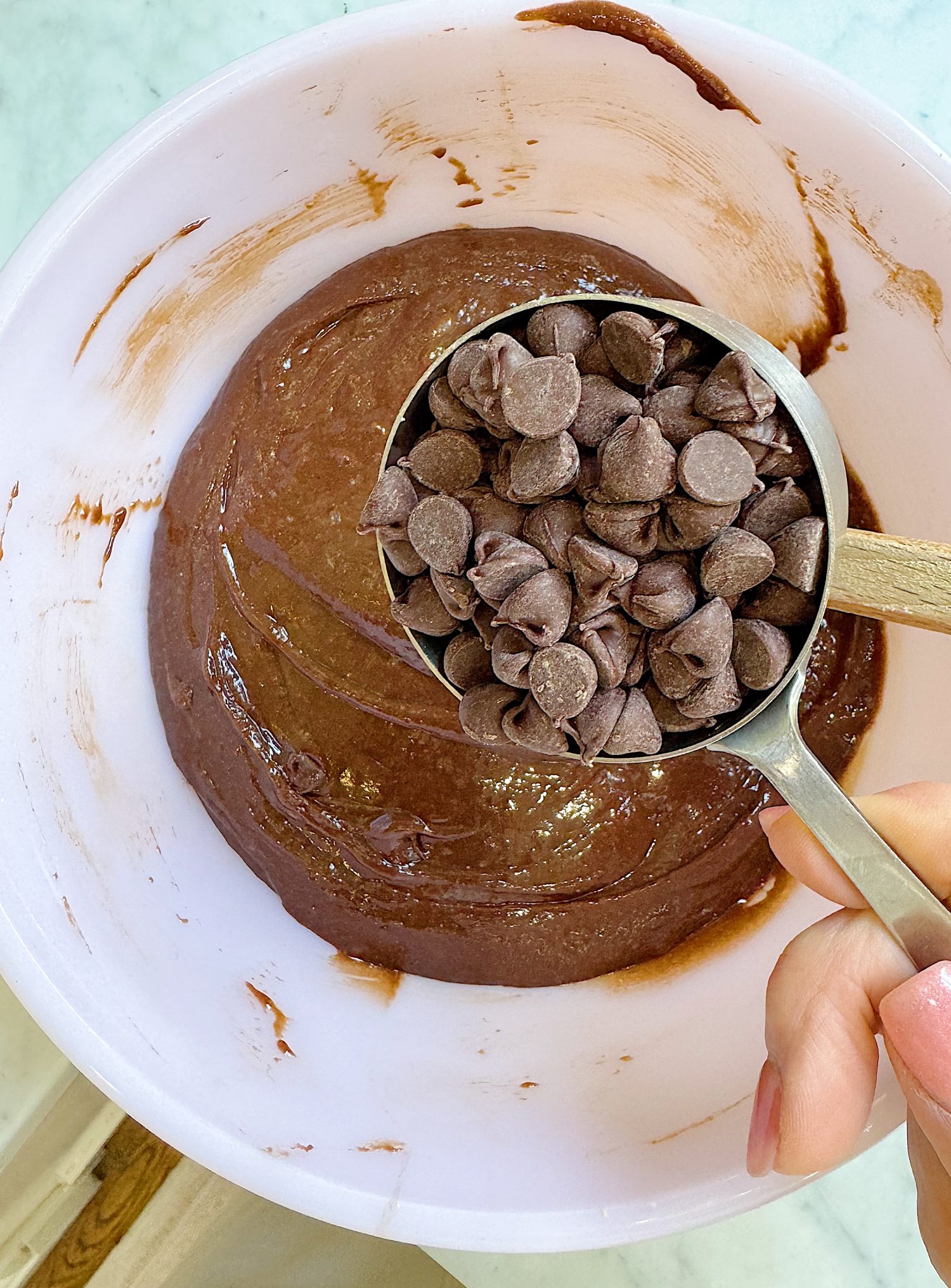 A hand holds a measuring cup filled with chocolate chips over a mixing bowl containing chocolate batter.