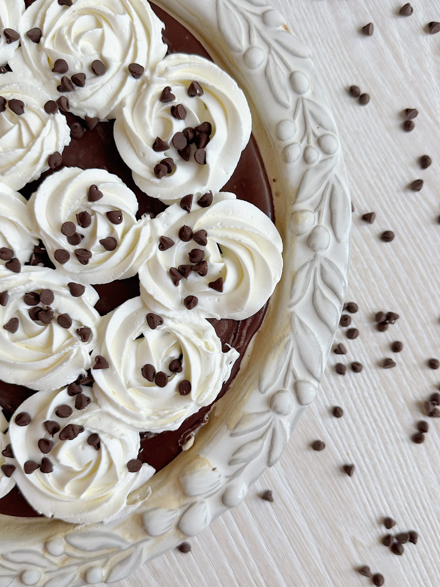 A pie topped with swirls of whipped cream and sprinkled with chocolate chips, displayed in a decorative white pie dish on a light wooden surface.