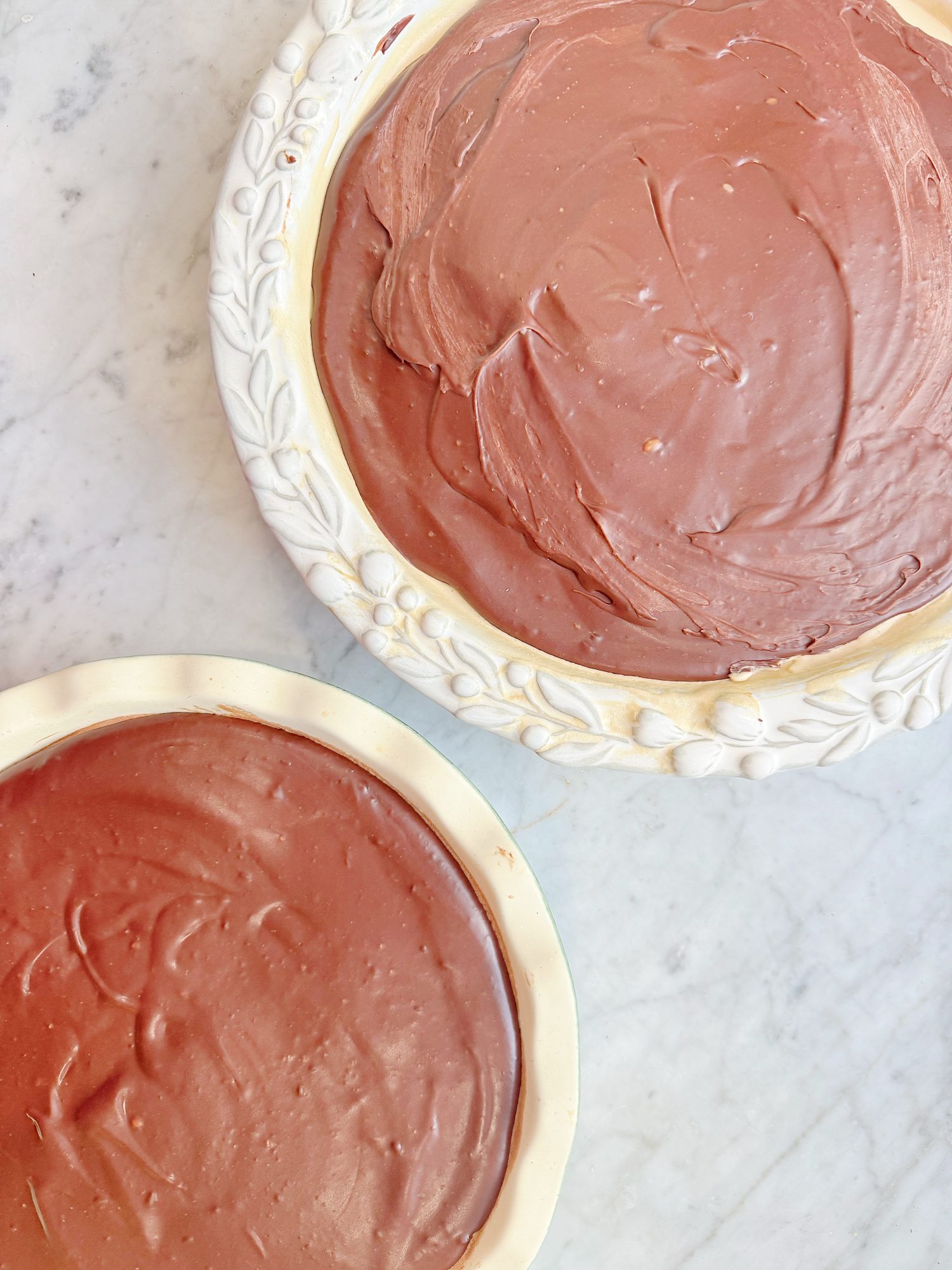 Two pie dishes filled with smooth, chocolate pie filling are placed on a light-colored, marble surface.