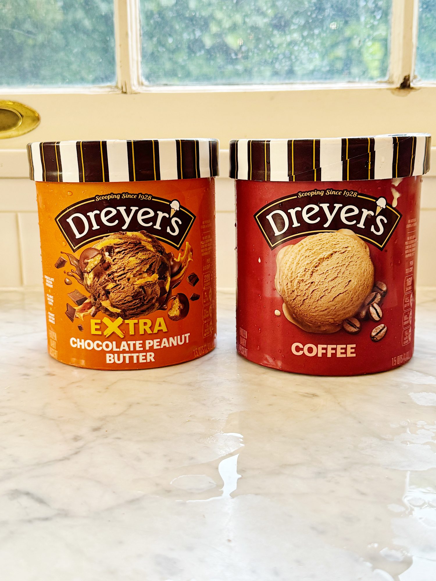 Two tubs of Dreyer's ice cream, one labeled "Extra Chocolate Peanut Butter" and the other labeled "Coffee," sit on a marble counter in front of a window.