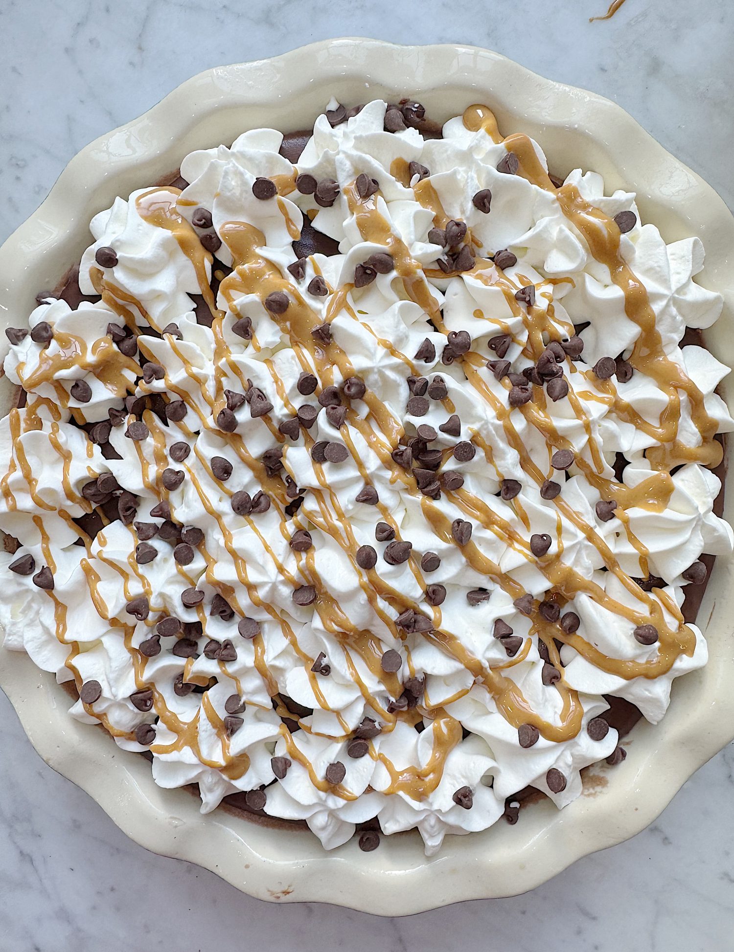 Pie topped with whipped cream, chocolate chips, and peanut butter drizzle in a white pie dish on a marble surface.