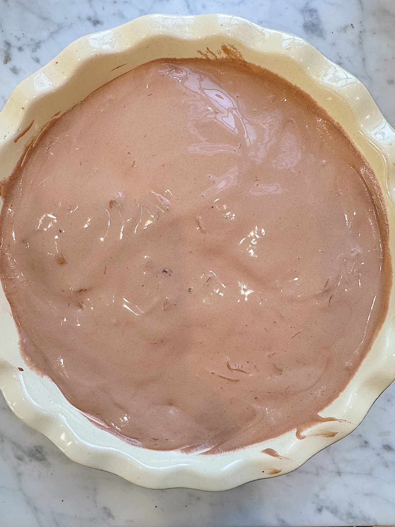A round pie dish filled with an even layer of smooth, light brown batter on a marble surface.