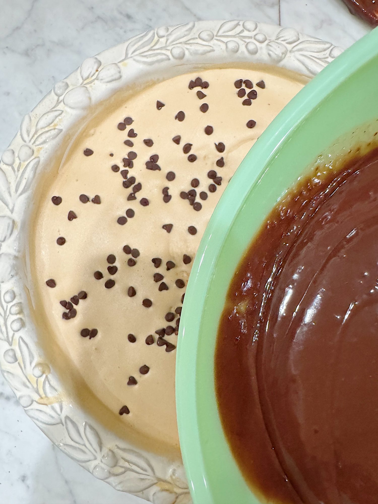 A pie pan with a light brown batter topped with chocolate chips sits next to a green bowl filled with smooth chocolate batter.