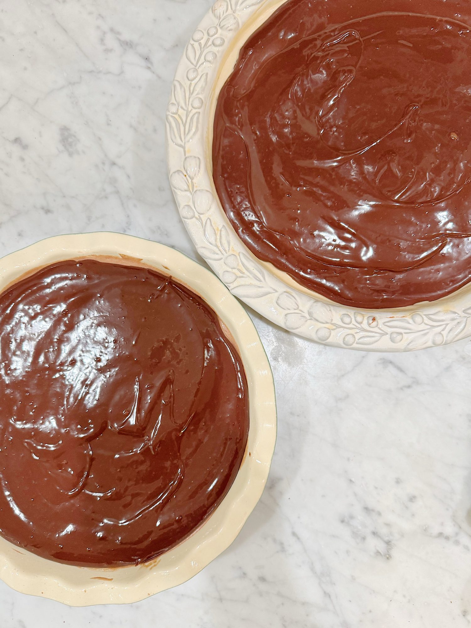 Two pie dishes filled with smooth, glossy chocolate pie filling sit on a white marble surface.