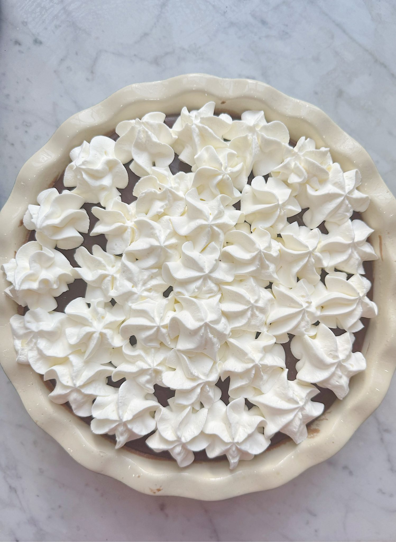 A chocolate pie in a white ceramic dish, topped with swirls of whipped cream, on a light marble surface.