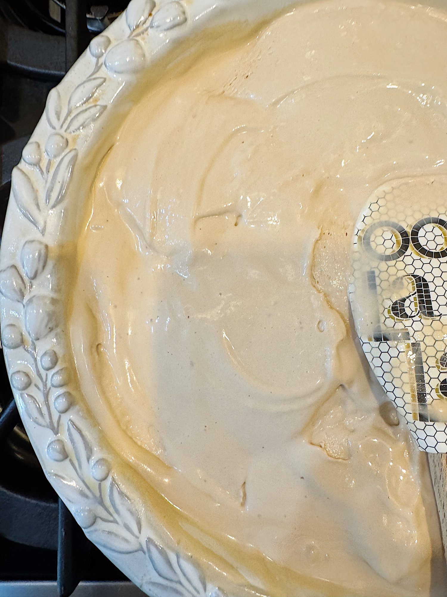 A ceramic pie dish filled with a smooth, pale batter being spread with a spatula, positioned on a stovetop.