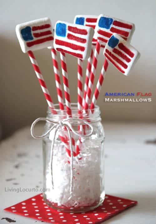 A mason jar holds red and white striped sticks with rectangular marshmallows decorated as American flags, set on a red polka dot napkin.