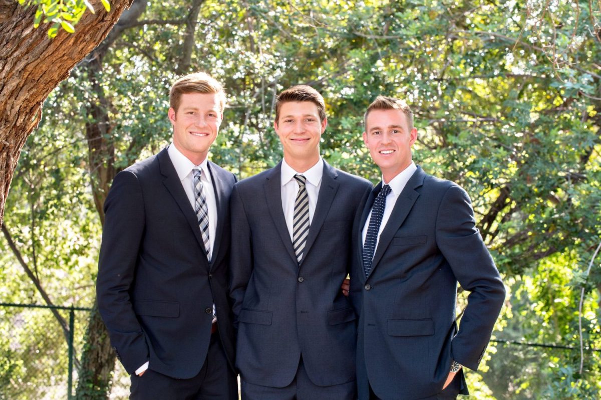 Three young men in suits stand outdoors in front of trees, posing together and smiling at the camera.