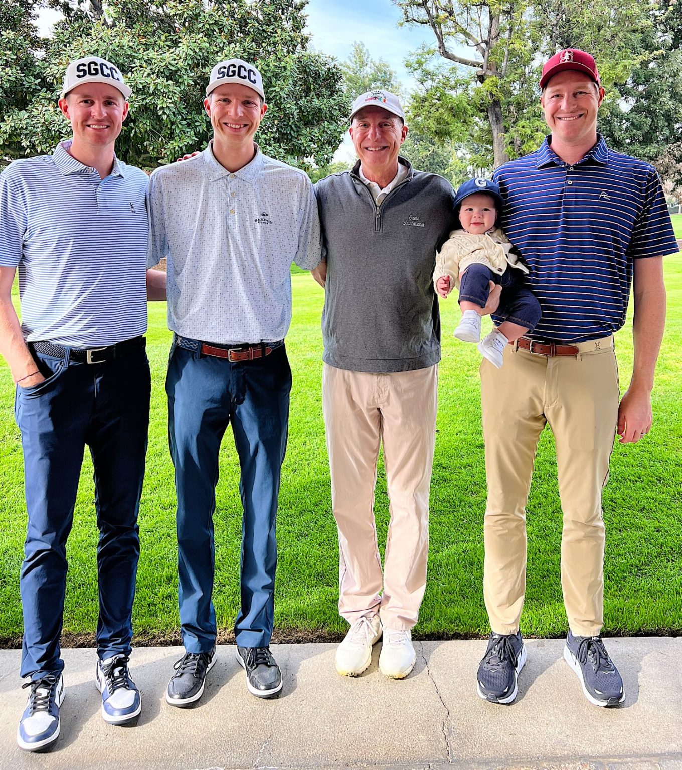 Four adults stand side by side outdoors on grass, with one holding a baby. All are dressed in golf attire and smiling at the camera.