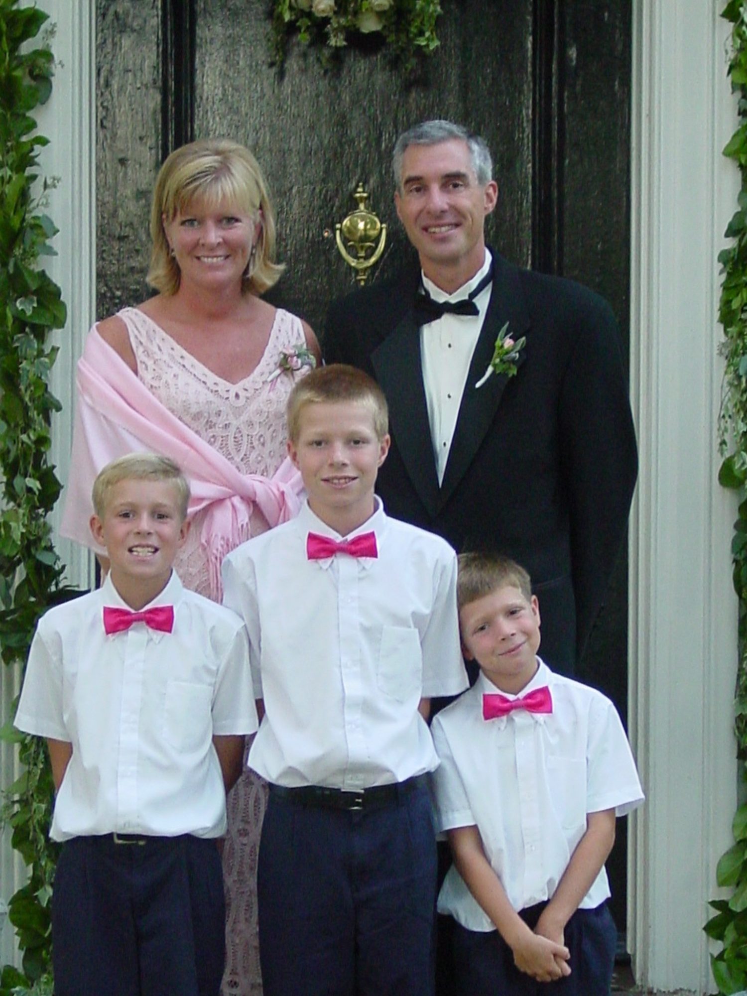 A woman in a pink dress and a man in a tuxedo stand behind three boys wearing white shirts and pink bow ties, posing for a photo in front of a black door decorated with greenery.