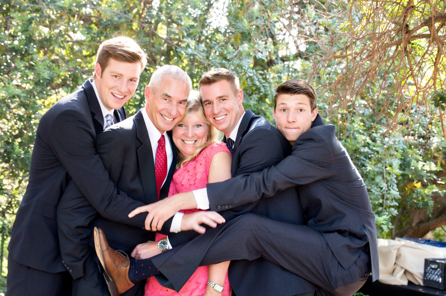 Four men in suits and a woman in a pink dress pose outdoors, smiling and hugging closely in front of green foliage on a sunny day.