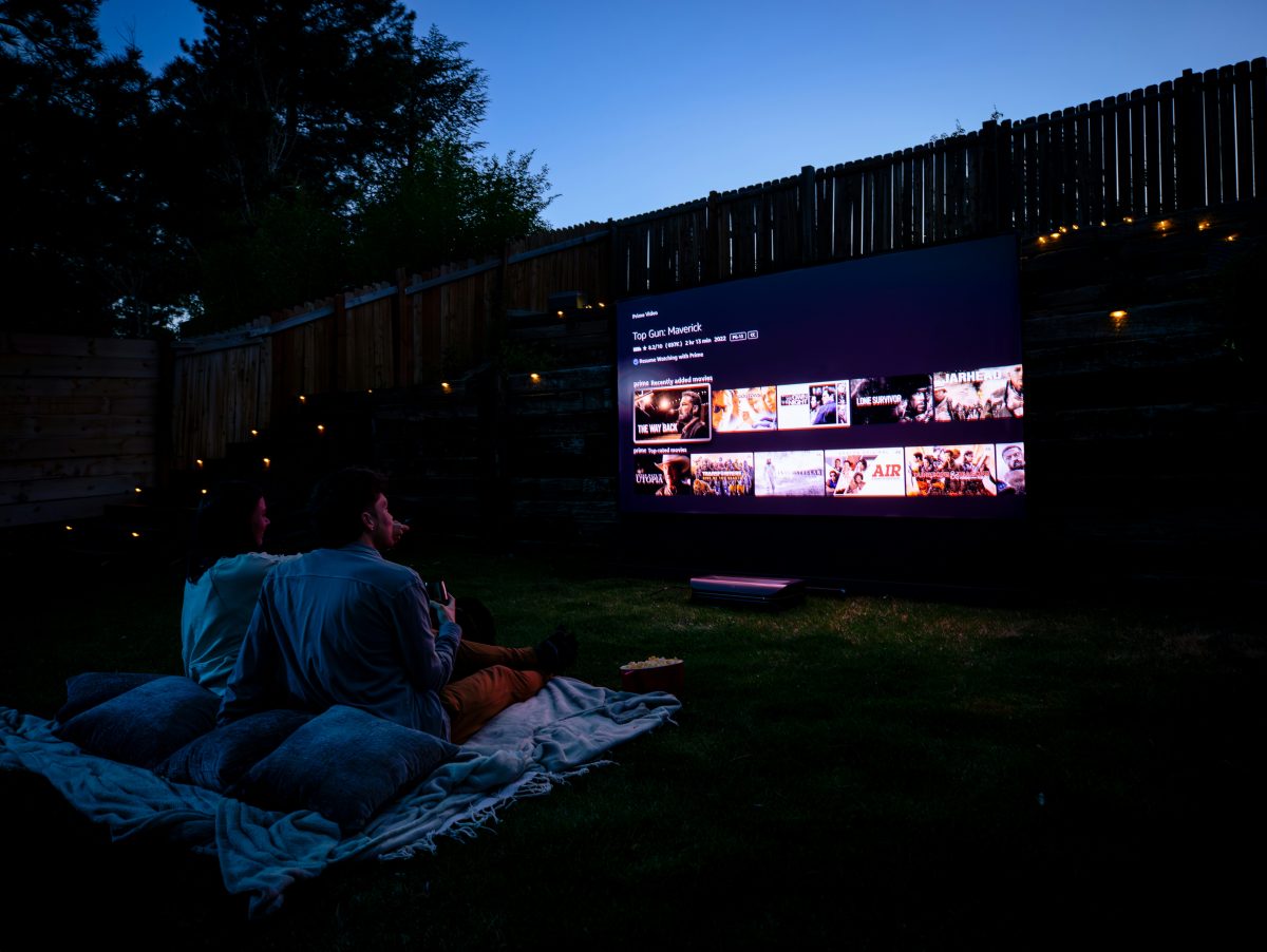 Two people sit on blankets in a backyard at dusk, watching a large outdoor screen displaying a streaming service menu.