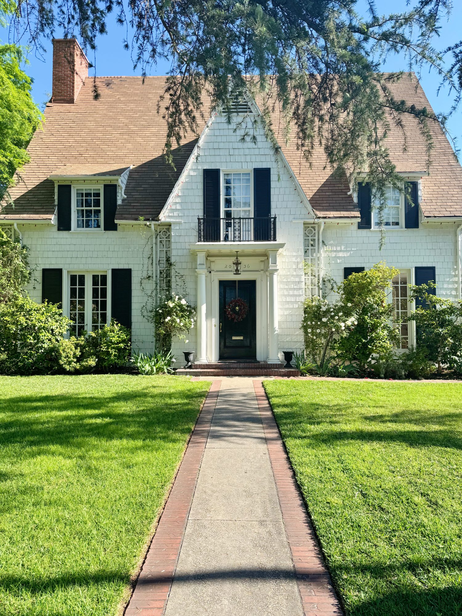 White two-story house with black shutters, steep gabled roof, central front door, and manicured lawn bordered by bushes and trees under a clear blue sky.
