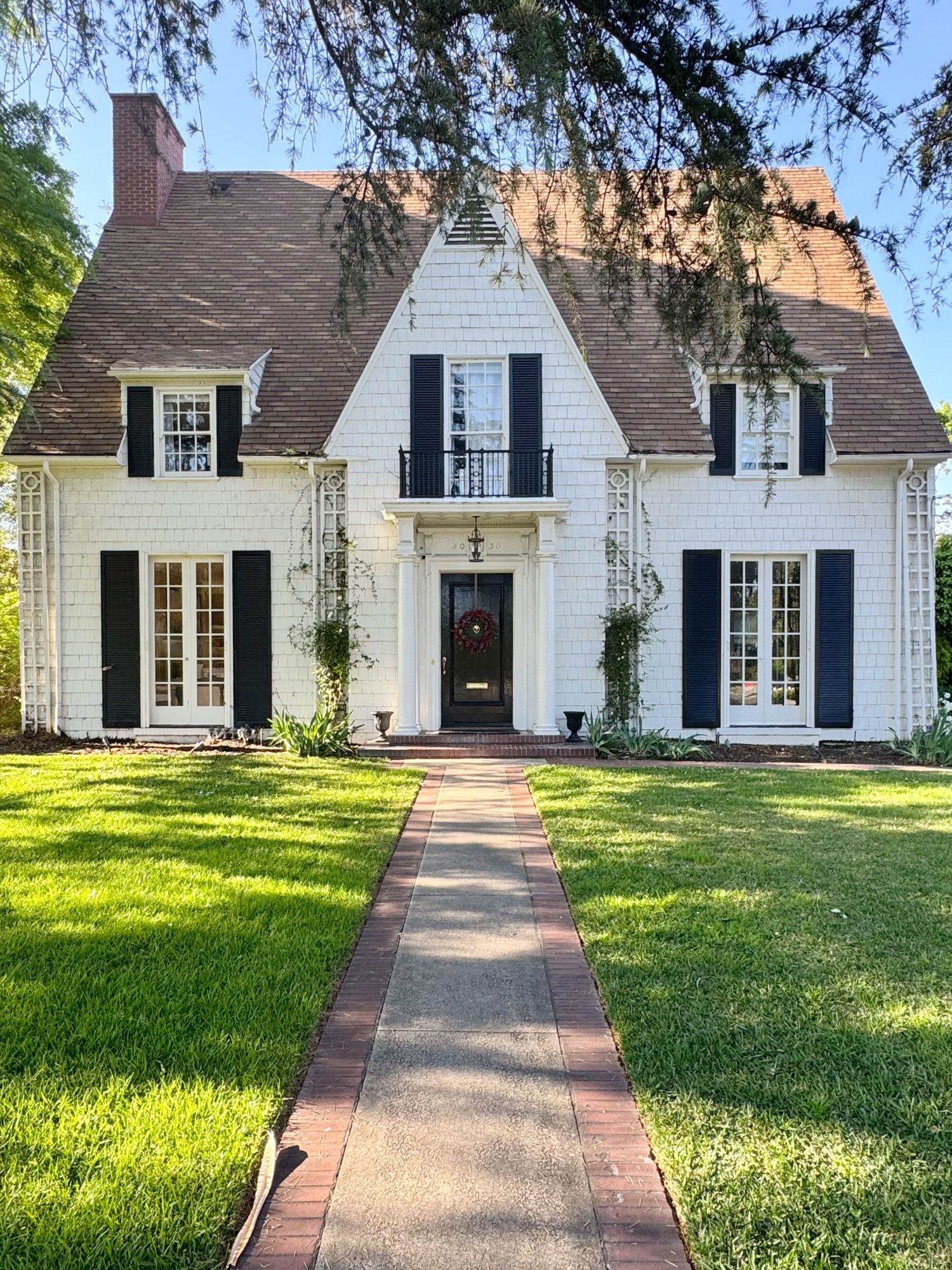 A two-story white house with dark shutters, a steep roof, central front door, second-story balcony, and a brick-bordered walkway leading through a well-kept lawn.