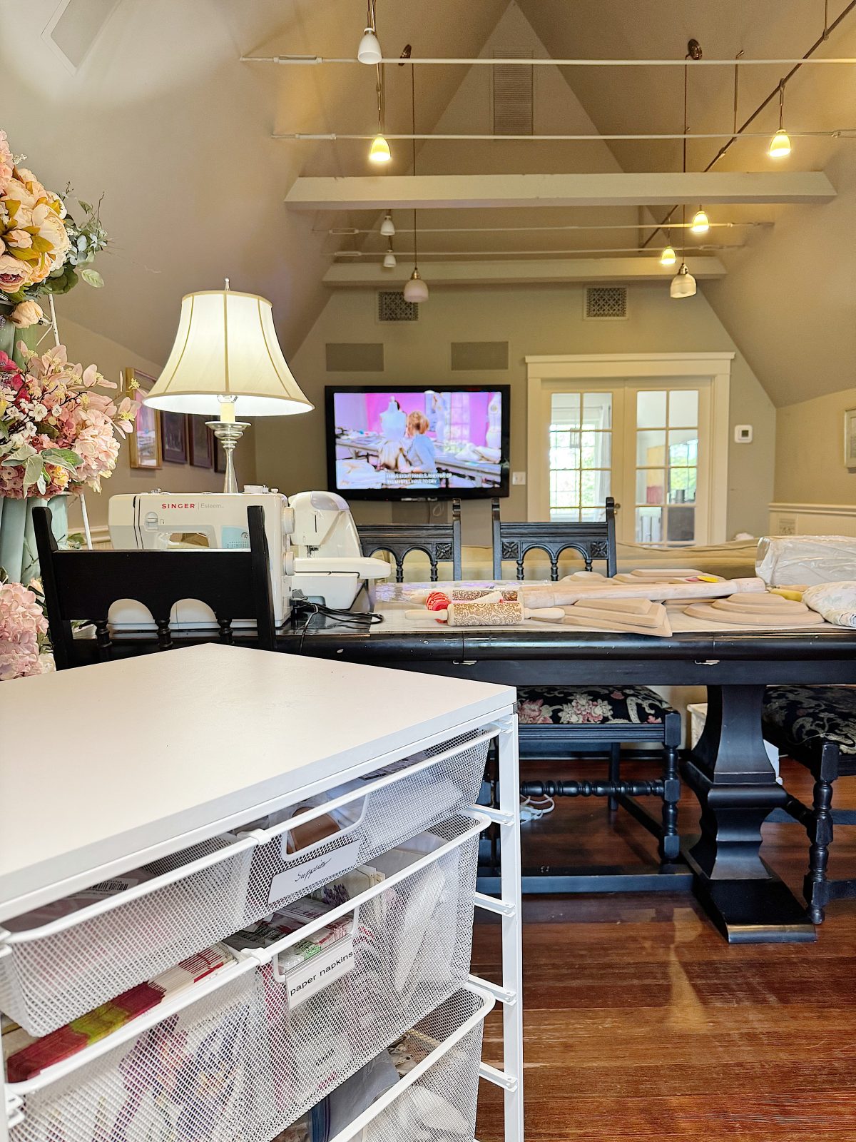 A sewing room with a large table, sewing machine, fabric, storage drawers, and a TV displaying a crafting show in the background.