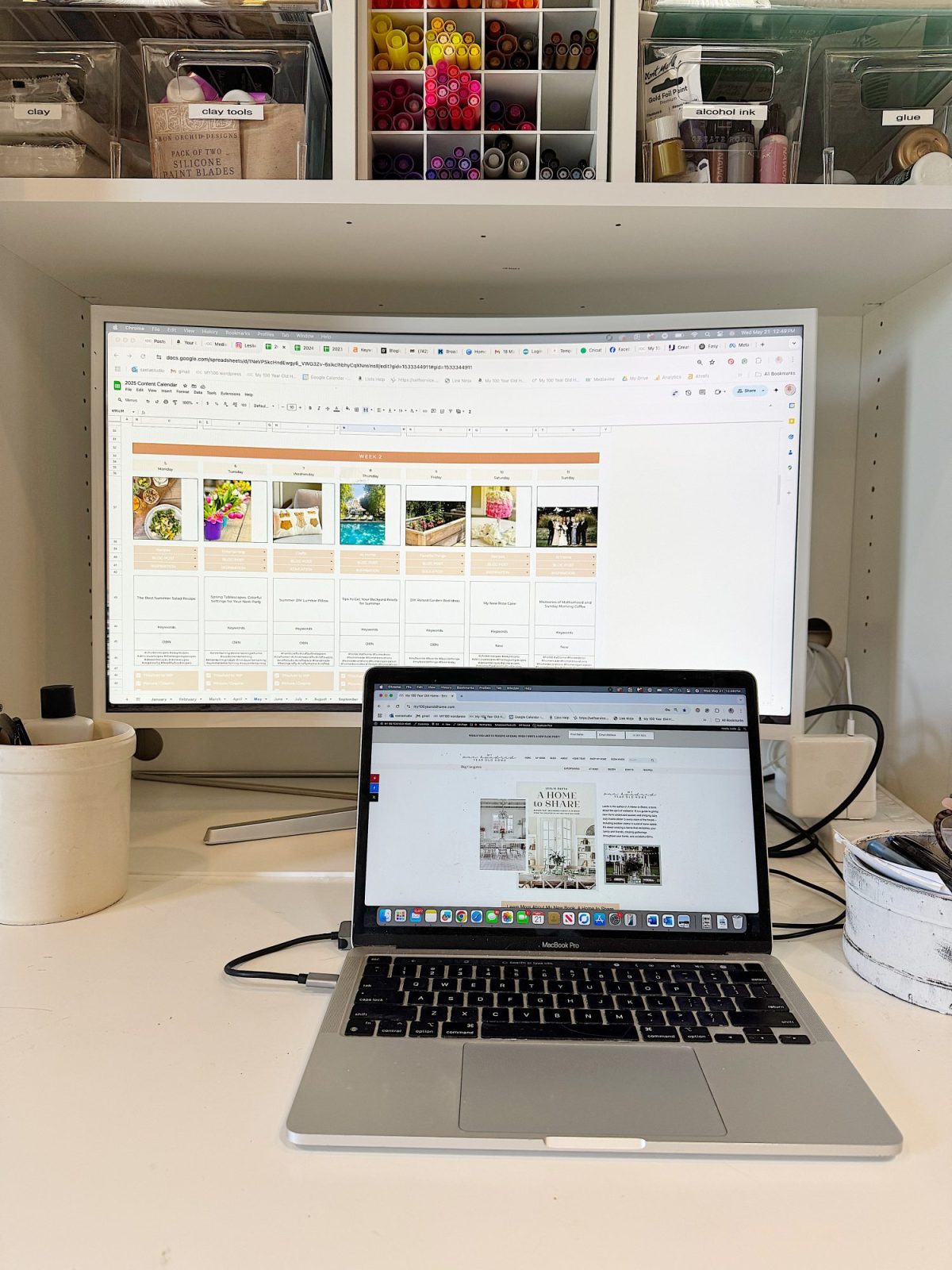 A workspace with a laptop in front of a large monitor displaying project boards, surrounded by craft supplies and organizers on a white desk.