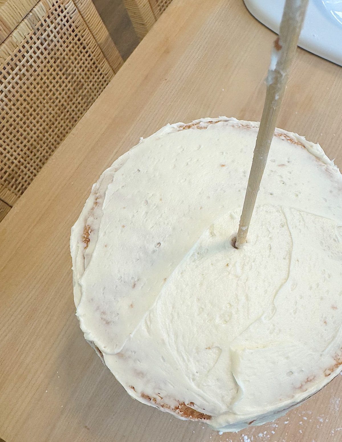 A round cake with light frosting sits on a wooden table, with a dowel inserted vertically through its center.