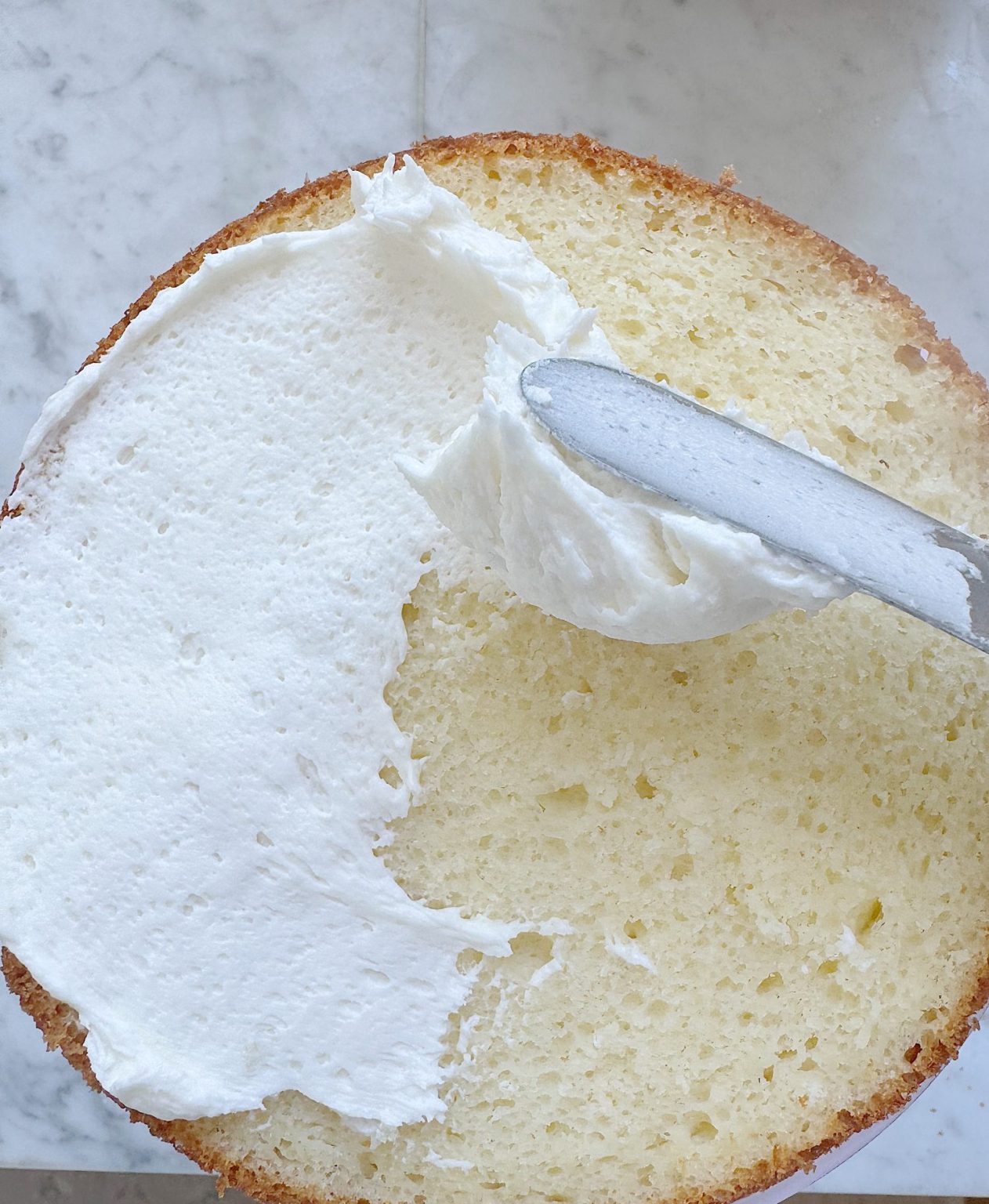A close-up of a cake being frosted with white icing using a metal spatula on a marble surface.