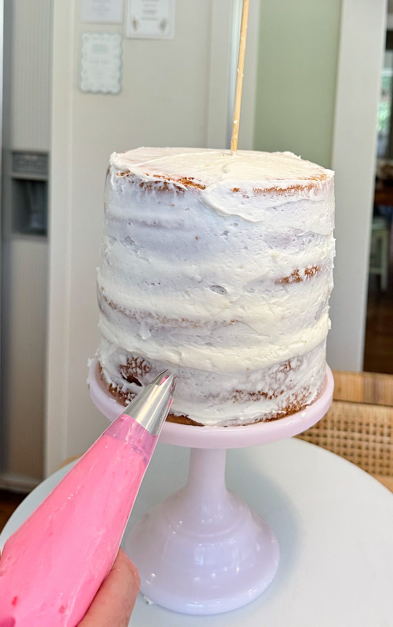 A partially frosted layer cake on a stand is being decorated with pink icing from a piping bag.