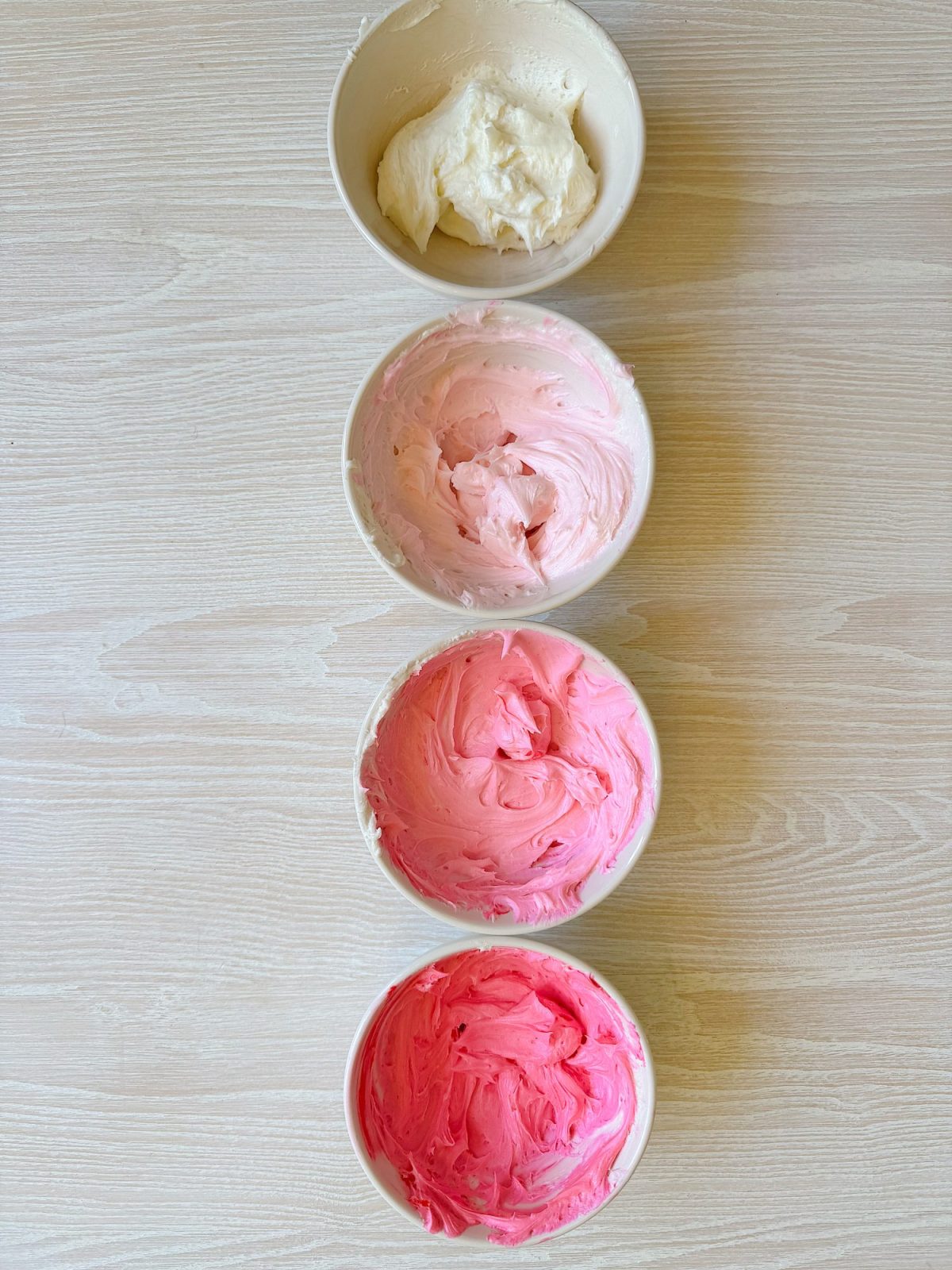 Four bowls of frosting arranged vertically on a light wooden surface, each showing a gradient from white at the top to deep pink at the bottom.