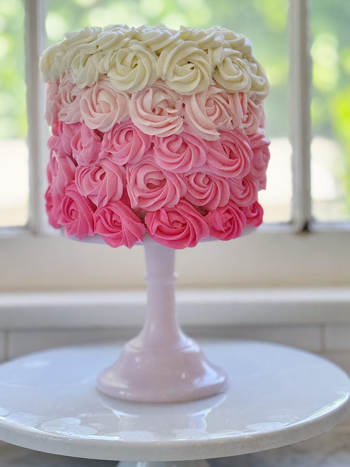 A round cake with pink and white rosette frosting sits on a white cake stand in front of a window.