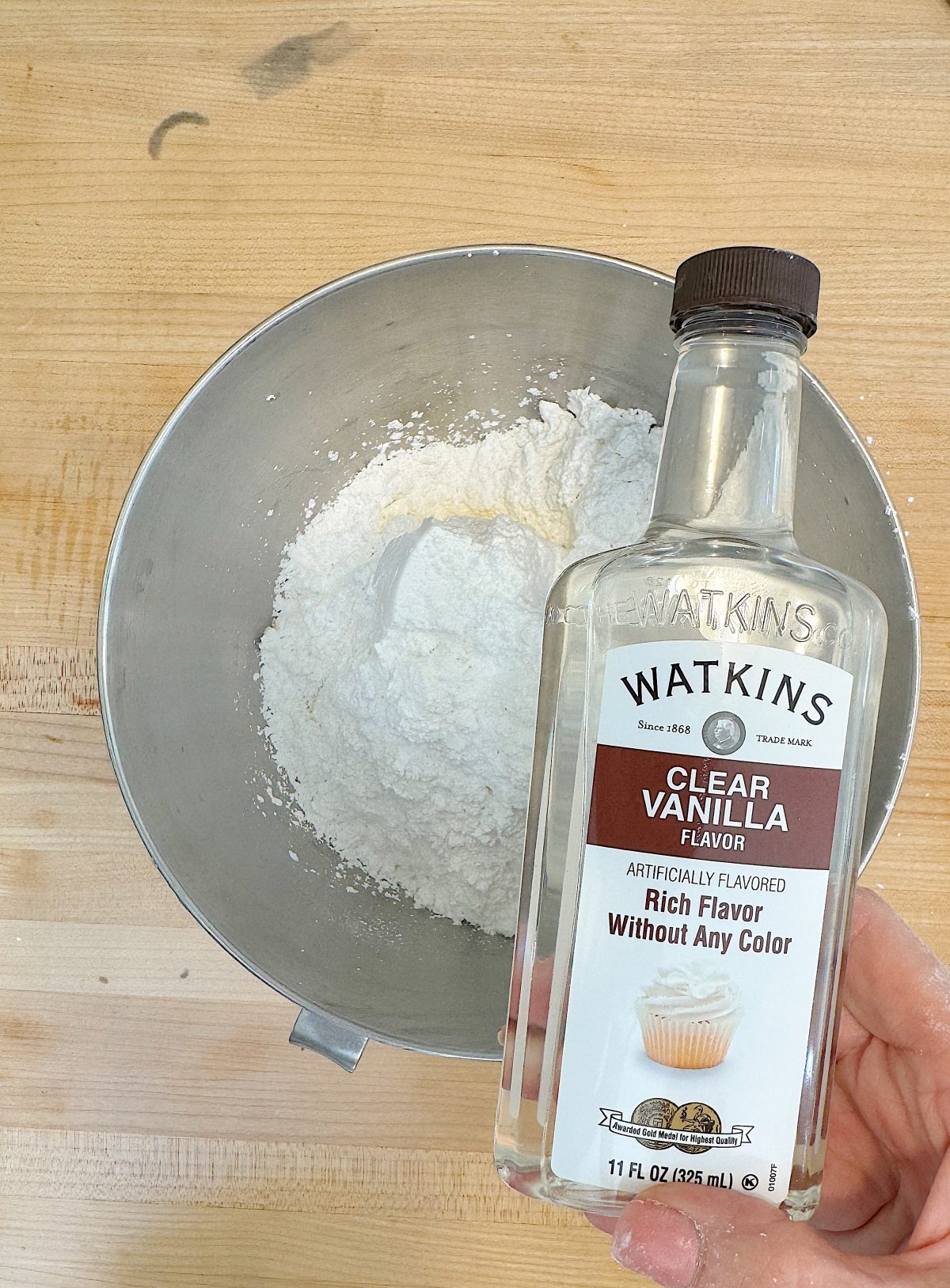 A hand holds a bottle of Watkins clear vanilla flavor above a metal bowl filled with flour on a wooden surface.