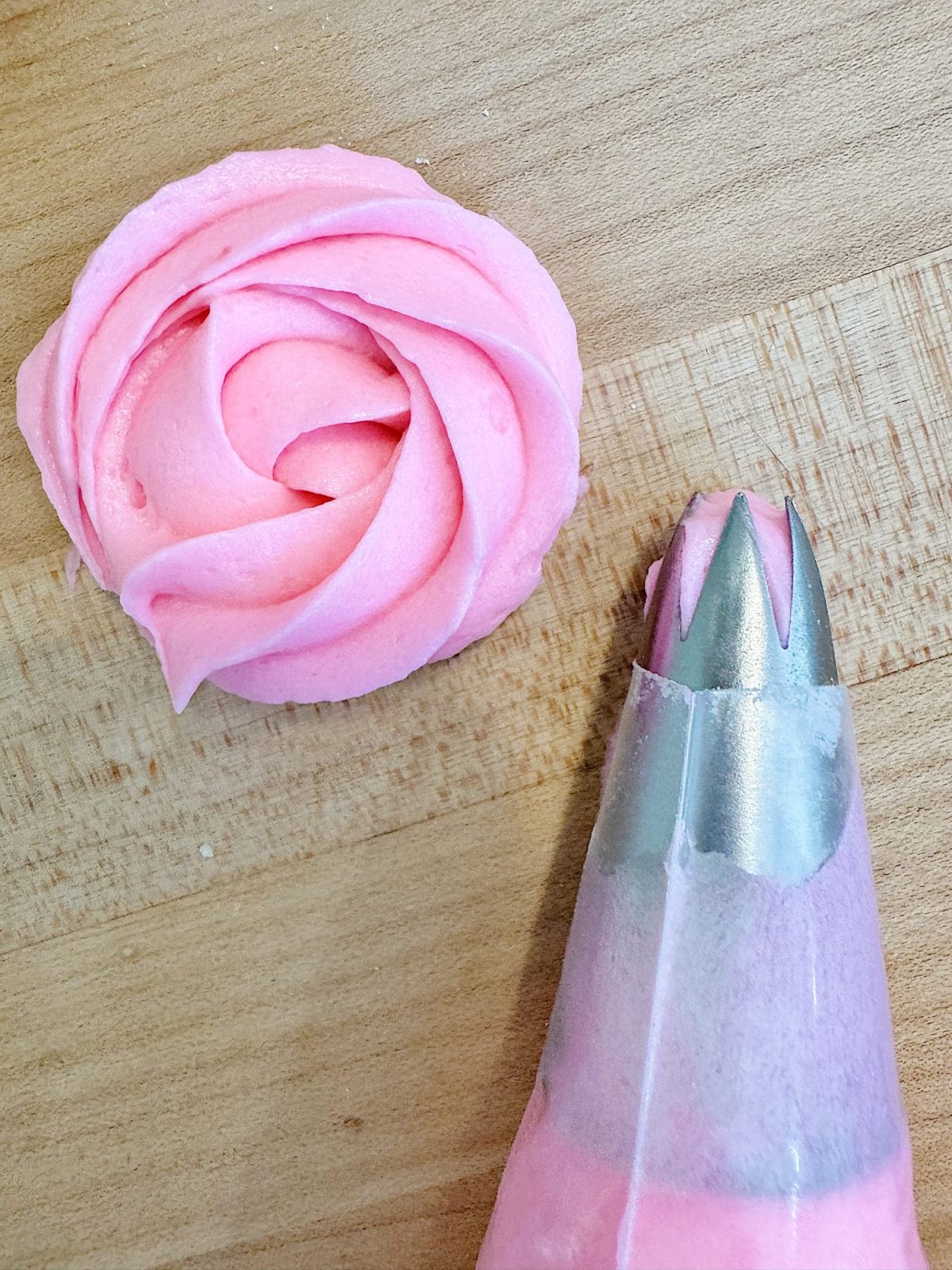A piping bag with pink frosting and a metal tip sits next to a pink buttercream rosette on a wooden surface.